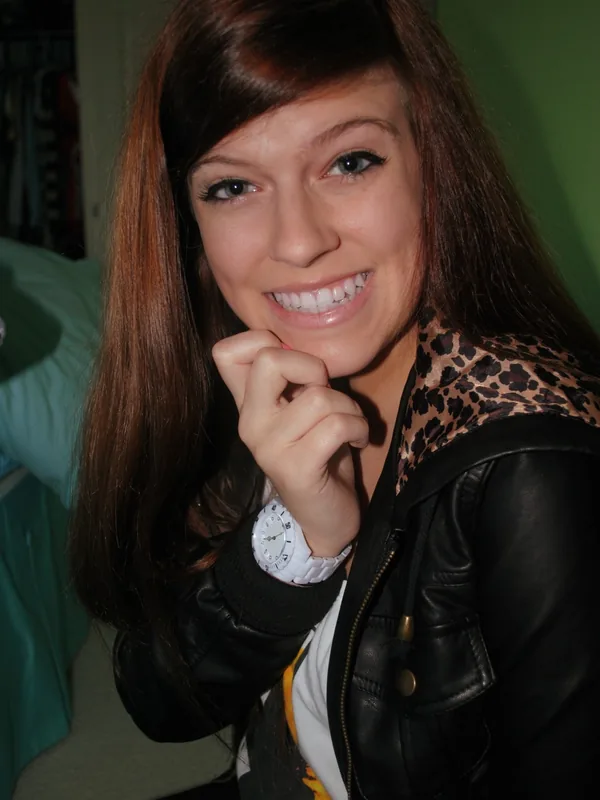 Portrait d'une jeune femme souriante aux cheveux bruns, portant une veste en cuir noire et une montre blanche, regardant l'objectif.