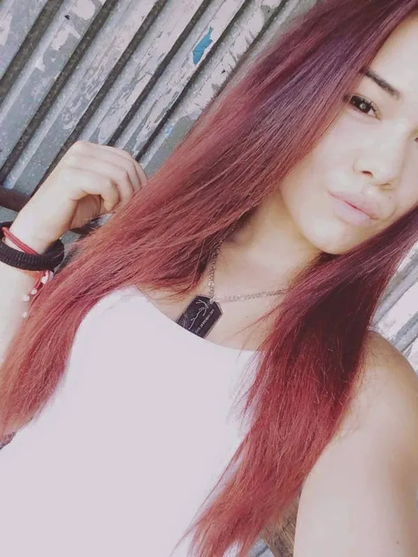 Portrait d'une jeune femme aux cheveux rouges portant un haut blanc, devant un mur en bois vieilli.