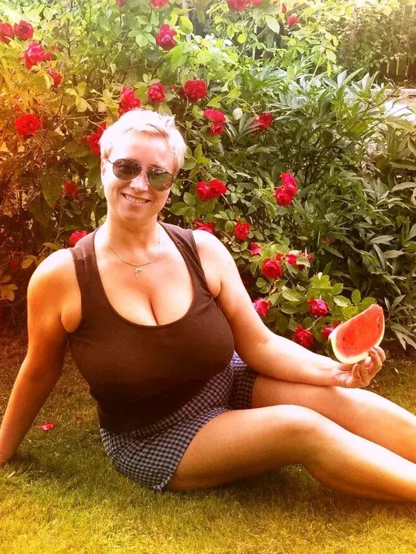 Femme aux cheveux courts assise sur l'herbe, tenant une tranche de pastèque devant des roses rouges.