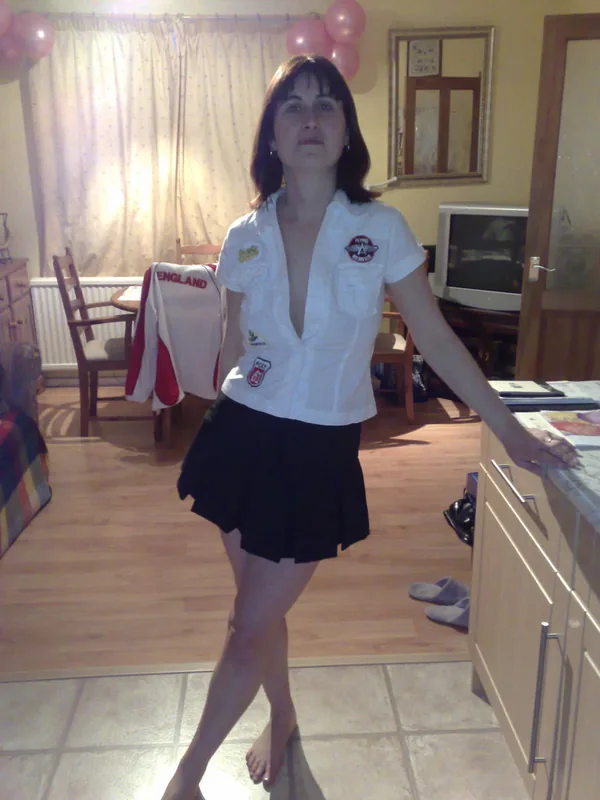 Femme debout dans un intérieur domestique, portant une chemise blanche avec badges et une jupe noire, pieds nus, regardant l'objectif.