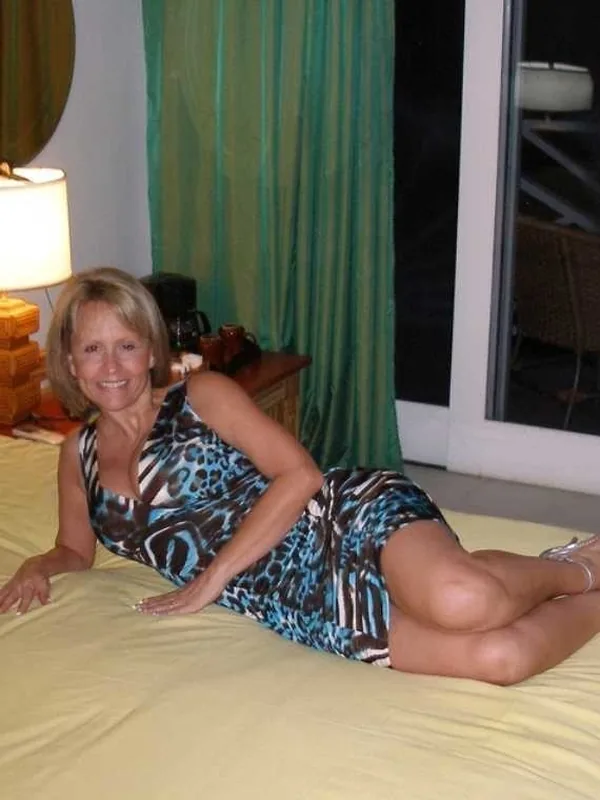 Femme blonde en robe imprimée allongée sur un lit jaune, souriante, dans une chambre éclairée par une lampe.