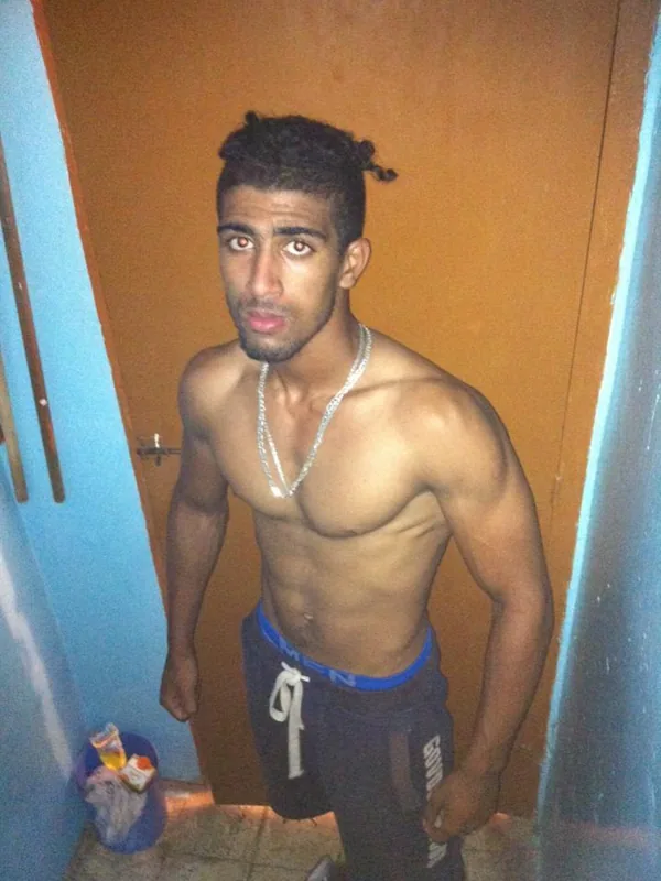 Homme torse nu portant un collier et des shorts, debout dans un couloir aux murs bleus et porte marron.