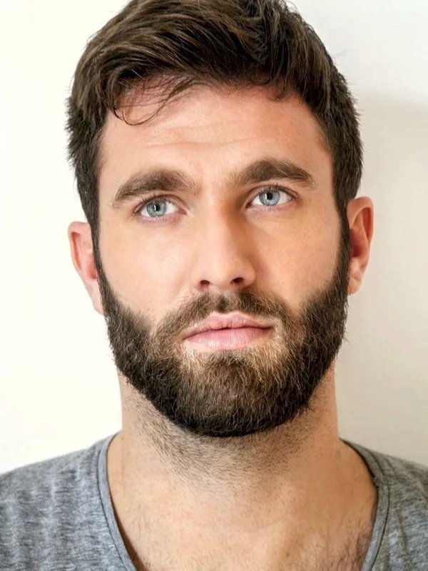Portrait en gros plan d'un homme aux cheveux bruns et barbe, portant un t-shirt gris, regardant l'objectif.