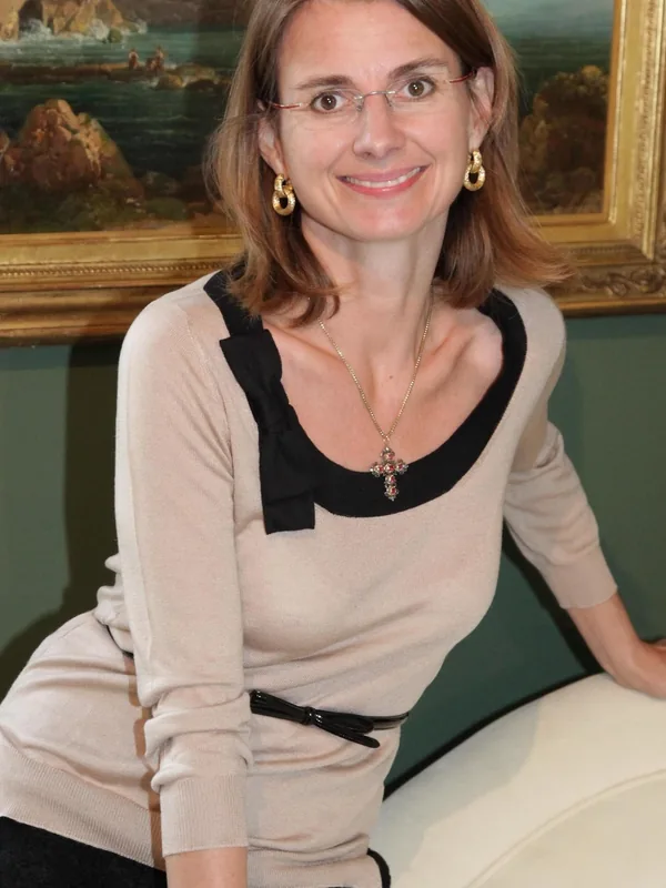 Portrait d'une femme aux cheveux bruns, portant des lunettes et un haut beige avec col noir, souriant devant un tableau marin.