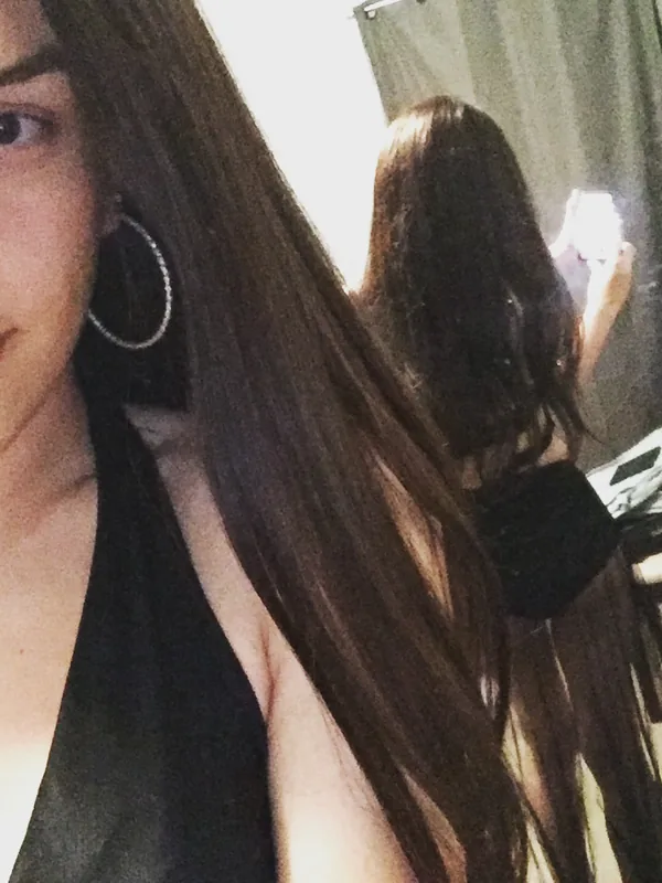 Portrait d'une femme aux longs cheveux bruns prenant un selfie devant un miroir, portant un haut noir et des boucles d'oreilles larges.