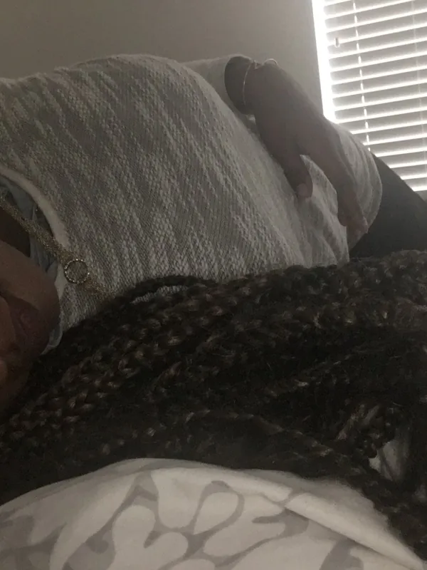 Femme allongée sur un lit, regardant l'objectif, cheveux tressés, vêtue d'un haut gris, lumière naturelle par des volets.