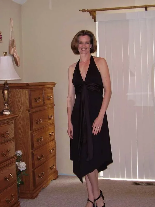 Femme en robe noire debout dans une chambre, devant un meuble en bois et une fenêtre aux stores verticaux.