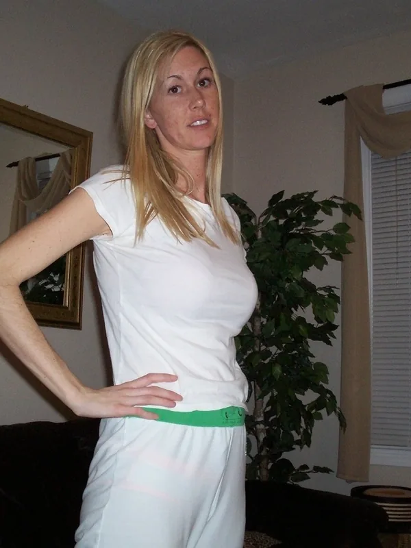 Femme blonde en tenue blanche avec ceinture verte, debout dans un salon, regardant l'objectif.