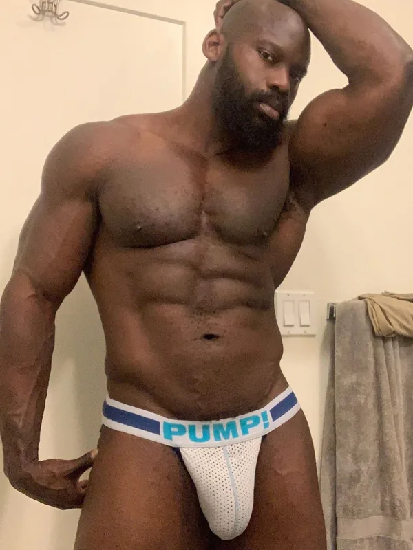 Homme noir musclé en string blanc avec inscription PUMP!, posant dans une pièce intérieure.