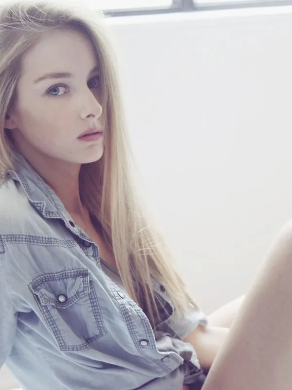 Jeune femme blonde assise, vêtue d'une chemise en jean, regardant l'objectif dans un intérieur lumineux.