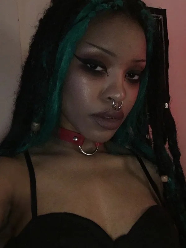 Portrait d'une femme aux cheveux tressés verts, portant un collier rouge et un haut noir, regardant l'objectif.