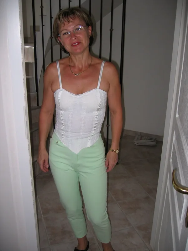 Femme aux cheveux courts portant des lunettes, un haut blanc à bretelles et un pantalon vert pâle, debout dans un couloir intérieur.