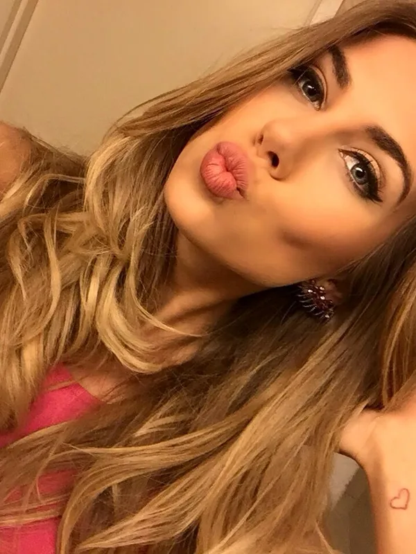 Portrait en gros plan d'une femme aux cheveux blonds ondulés faisant une moue, portant un haut rose et regardant l'objectif.