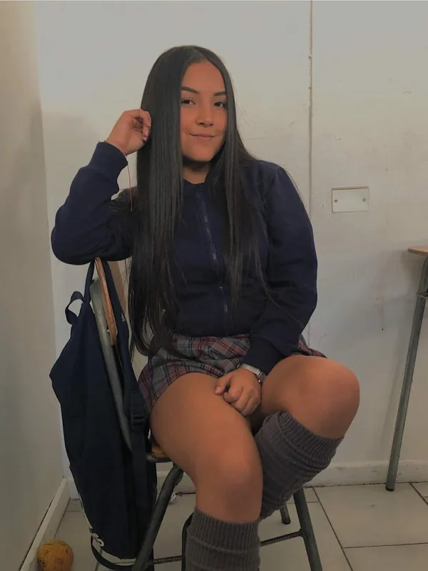 Jeune femme assise sur une chaise, vêtue d'un sweat bleu, d'une jupe à carreaux et de chaussettes hautes, regardant l'objectif.