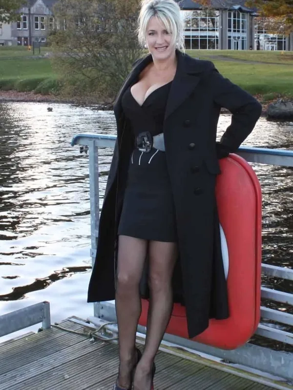 Femme aux cheveux blonds portant une robe noire et un manteau, posant sur un ponton près d'un lac avec un gilet de sauvetage rouge.