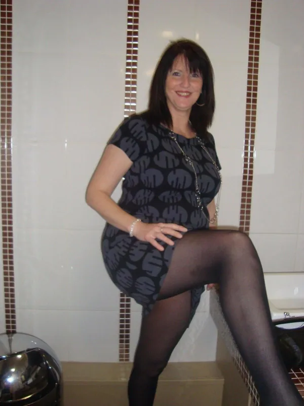 Femme assise sur le rebord d'une baignoire, vêtue d'une robe à motifs et de collants noirs, dans une salle de bain carrelée.