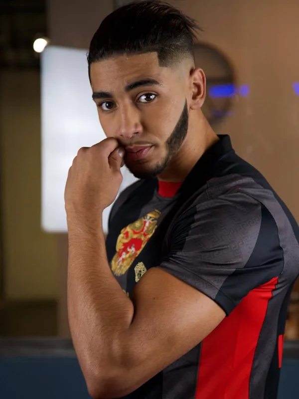 Portrait d'un homme aux cheveux courts et barbe, portant un t-shirt noir et rouge, regardant l'objectif avec la main près du visage.