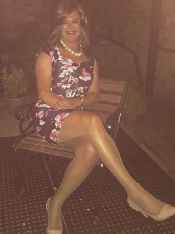 Femme assise sur un banc en extérieur la nuit, portant une robe fleurie, des collants et des escarpins, souriant vers l'objectif.