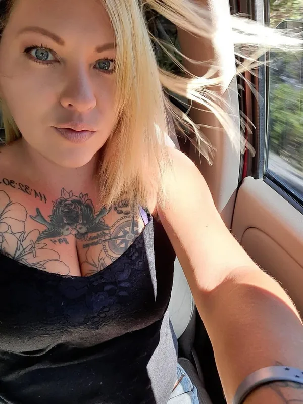 Portrait d'une femme blonde aux yeux bleus, tatouée, portant un haut noir en dentelle, assise dans une voiture avec la fenêtre ouverte.