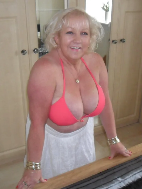 Femme blonde en haut de bikini rose et jupe blanche, appuyée sur un lit dans une chambre lumineuse.