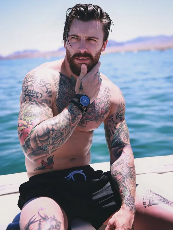 Homme tatoué torse nu assis sur un bateau, regardant l'objectif, avec une étendue d'eau et des montagnes en arrière-plan.