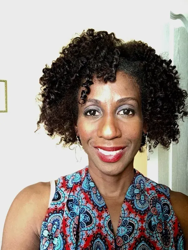 Portrait en buste d'une femme souriante aux cheveux bouclés, portant un haut coloré à motifs, regardant l'objectif.