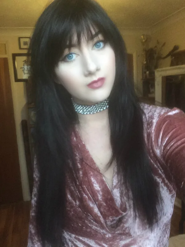 Portrait d'une femme aux cheveux noirs et yeux bleus, portant une robe rose et un collier strassé, dans un intérieur éclairé.