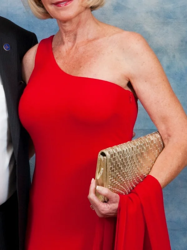 Femme aux cheveux blonds courts portant une robe rouge asymétrique, tenant un sac doré, posant devant un fond bleu.