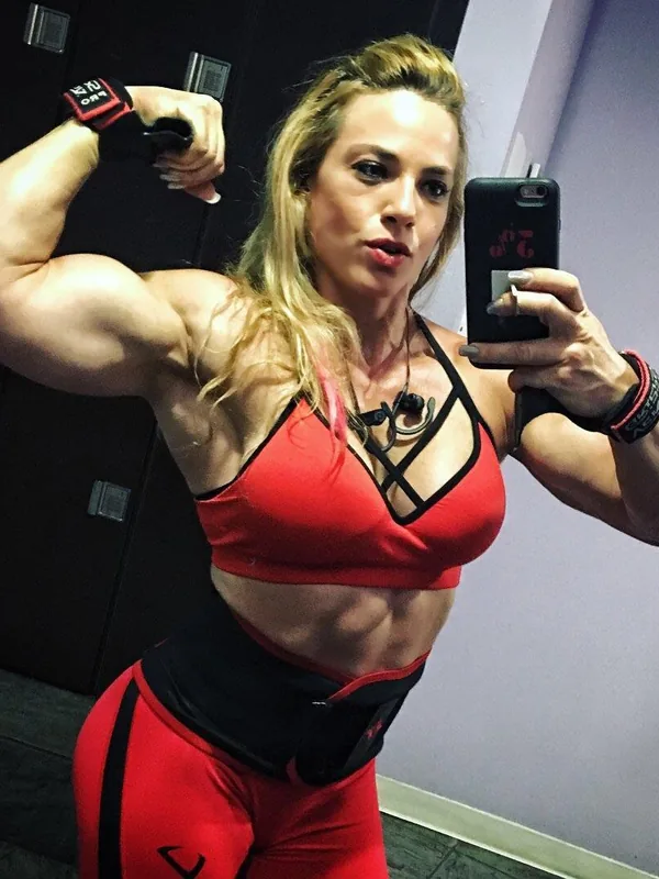 Femme blonde en tenue de sport rouge prenant un selfie dans un miroir, montrant ses muscles du bras et du ventre.