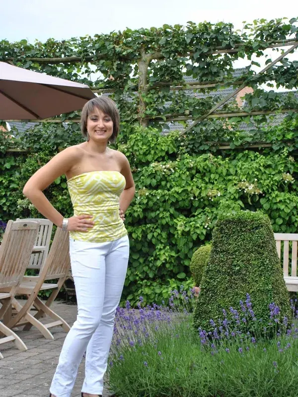 Femme souriante en tenue d'été posant dans un jardin avec des plantes vertes et des fleurs violettes.