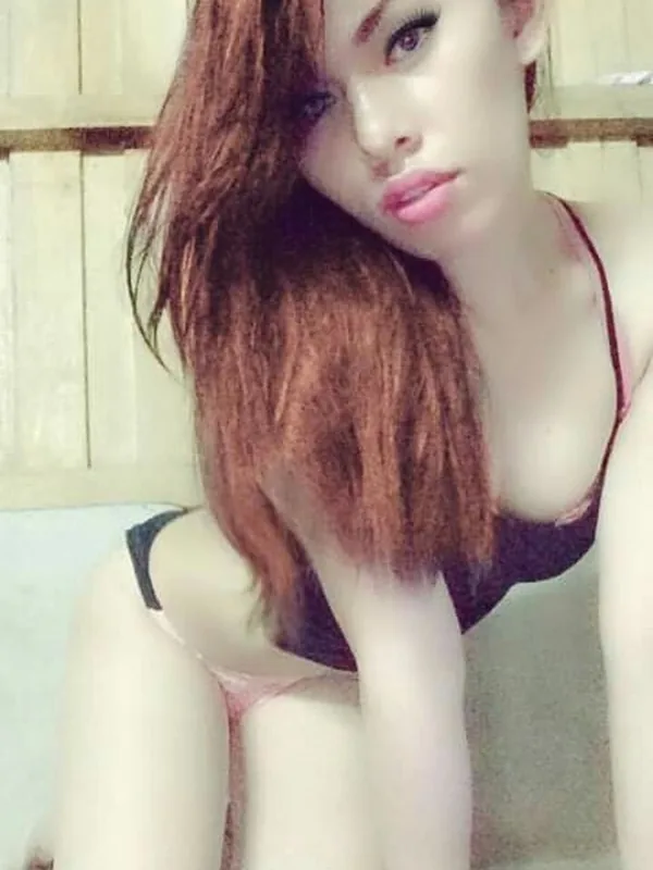 Femme aux cheveux auburn portant un haut de lingerie noir, assise sur un lit, regardant l'objectif.