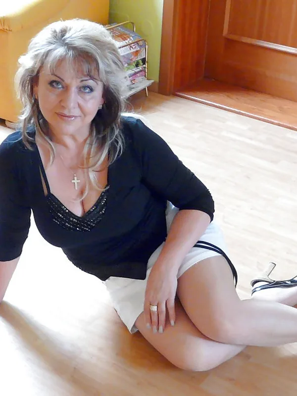 Femme aux cheveux blonds assise sur un sol en bois, vêtue d'une chemise noire et d'une jupe blanche, regardant l'objectif.