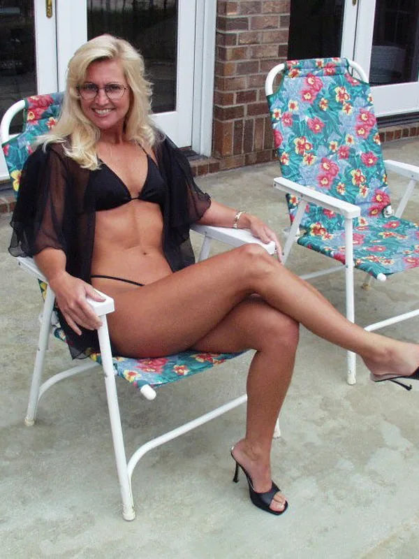 Femme blonde en bikini noir et veste transparente assise sur une chaise pliante fleurie sur une terrasse.