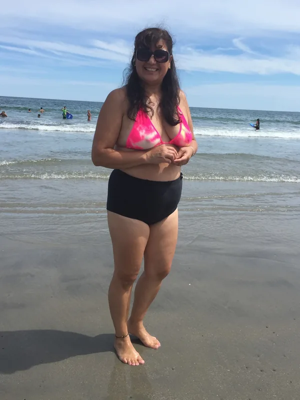 Femme en maillot de bain rose et noir sur une plage, souriante, pieds nus dans le sable humide près de l'eau.