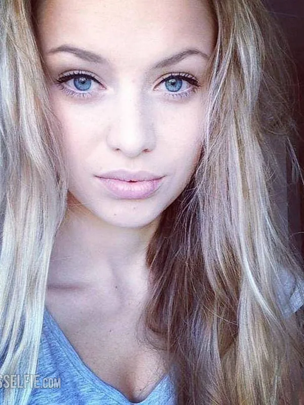Portrait en gros plan d'une jeune femme aux cheveux blonds et yeux bleus, portant un haut bleu clair, regardant l'objectif.