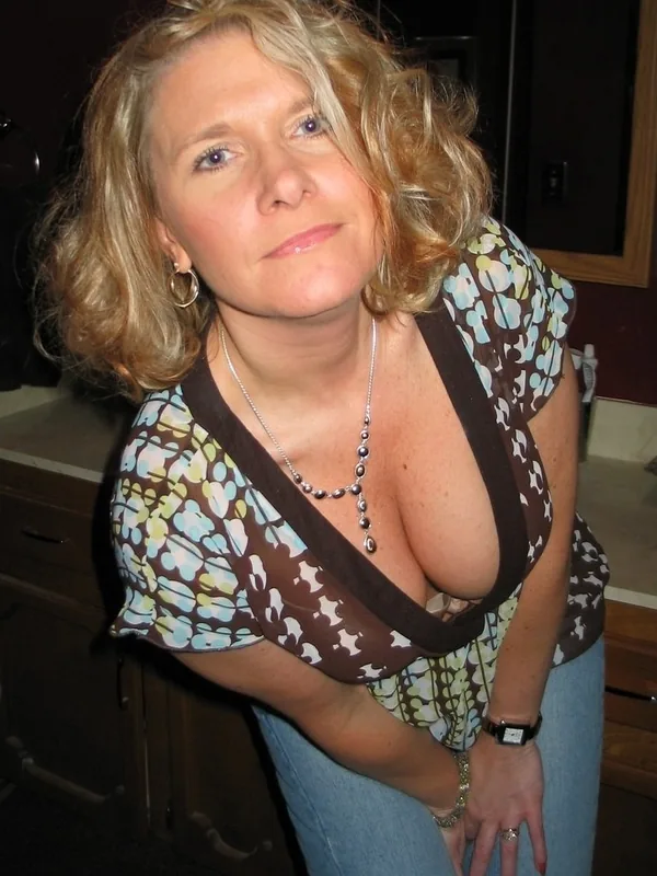 Portrait d'une femme aux cheveux blonds ondulés portant un haut à motifs floraux et un décolleté profond, dans un cadre intérieur.
