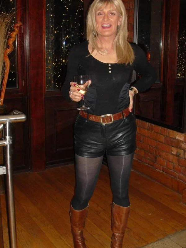 Femme blonde souriante en tenue noire tenant un verre de vin, debout dans un intérieur chaleureux avec parquet et briques.