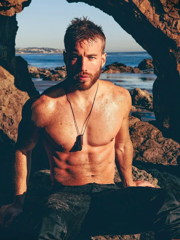 Homme torse nu assis sous une arche rocheuse au bord de la mer, regardant l'objectif.