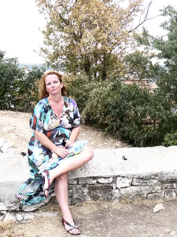 Femme aux cheveux roux en robe colorée assise sur un mur de pierre en extérieur.