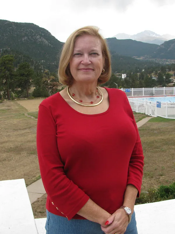 Femme aux cheveux blonds portant un haut rouge, debout devant un paysage montagneux avec une piscine en arrière-plan.