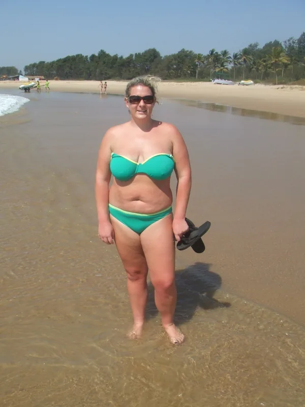 Femme en maillot de bain vert sur une plage, debout dans l'eau peu profonde, tenant des sandales.