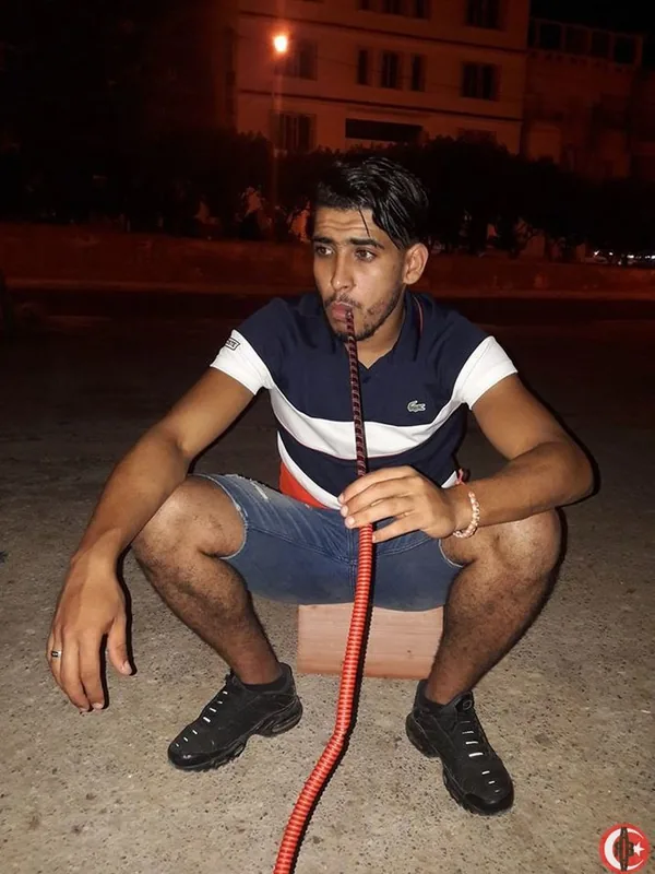 Homme assis sur un bloc de bois la nuit, tenant une pipe à eau rouge, vêtu d'un t-shirt Lacoste et de shorts en jean.