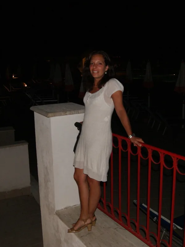 Femme en robe blanche souriante, appuyée sur une rambarde rouge sur un balcon la nuit.