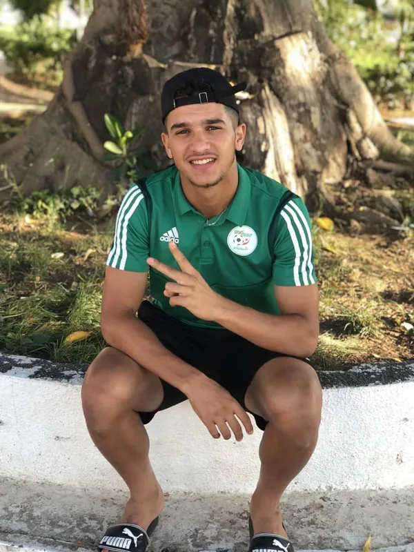 Jeune homme souriant, assis sur un rebord, portant un polo vert Adidas et une casquette noire, faisant un signe de la main devant un arbre.