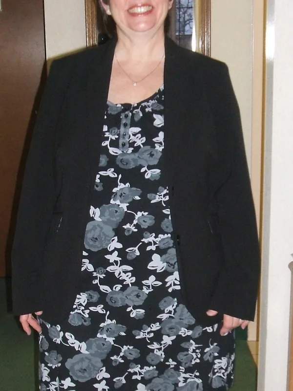 Portrait en pied d'une femme souriante portant une robe fleurie et un blazer noir, debout dans un intérieur éclairé.