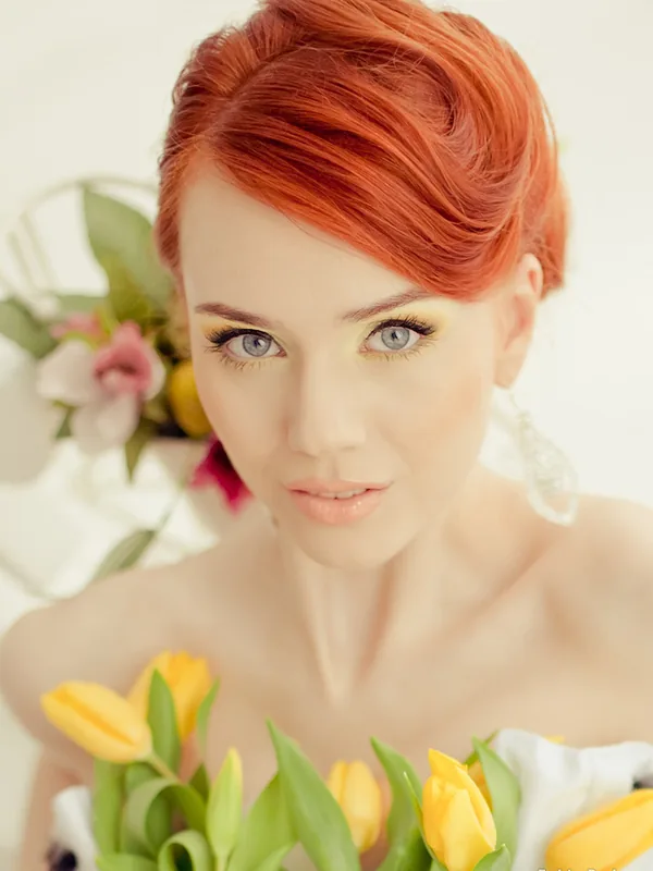 Portrait d'une femme aux cheveux roux et yeux bleus, tenant des tulipes jaunes, regardant l'objectif dans une lumière douce.