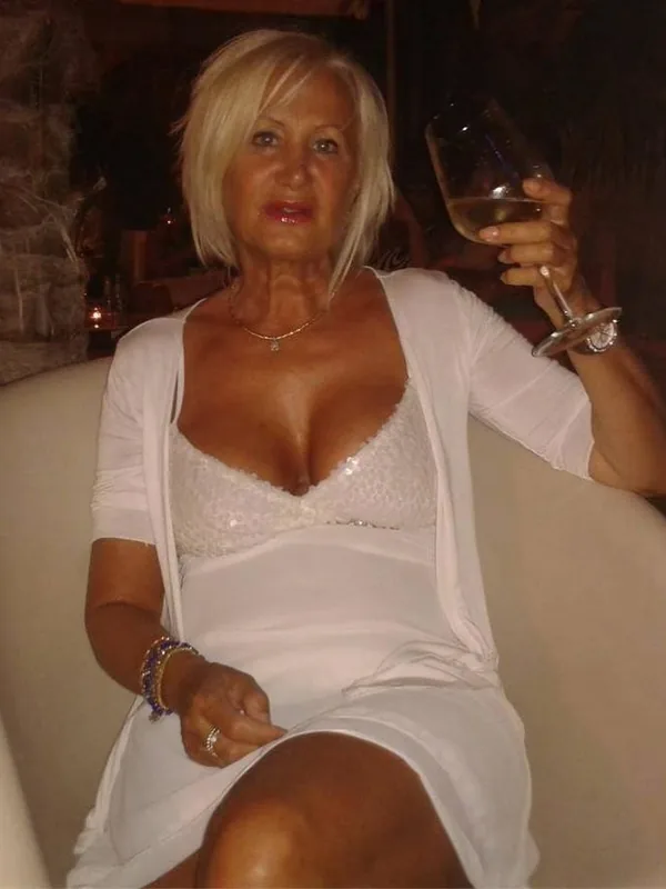 Femme blonde assise sur un canapé, portant une robe blanche et tenant un verre de vin blanc.