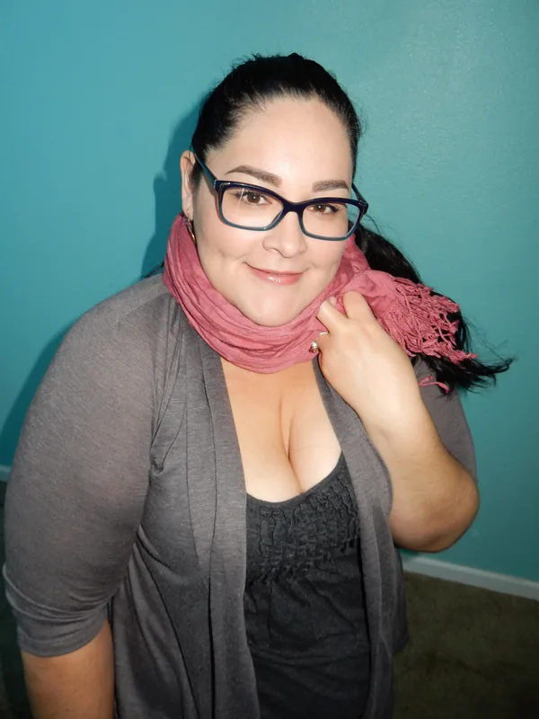 Femme aux cheveux noirs attachés, lunettes, foulard rose, cardigan gris, devant mur turquoise.