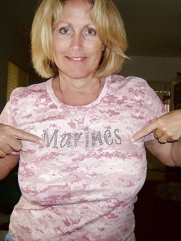 Femme blonde portant un t-shirt rose camouflage avec l'inscription Marines, pointant le texte avec les deux index.