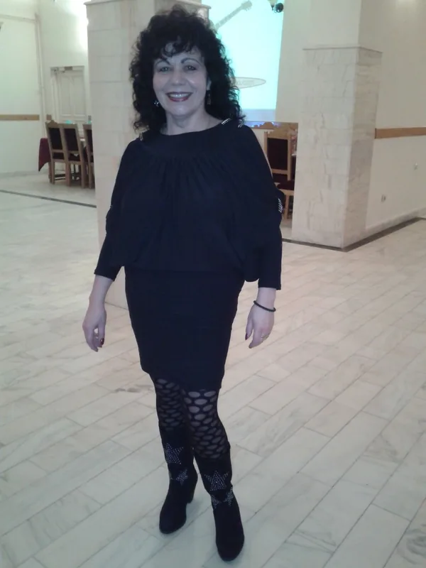 Femme souriante en tenue noire complète, debout dans un hall intérieur avec sol carrelé et colonnes.